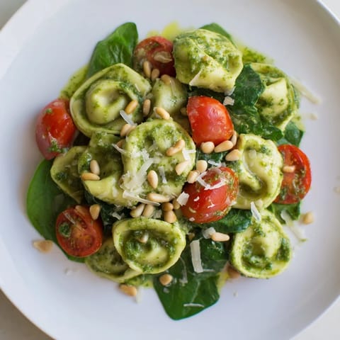 Tortellini salad with bright green pesto and red tomatoes, garnished with pine nuts and Parmesan, served on a platter.